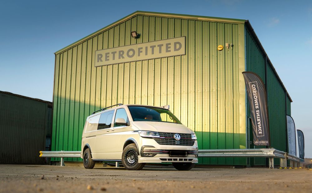 Cream vw campervan parked outside a large green building with Retrofitted sign above 